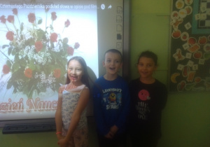 Uczeń i dwie uczennice spiewają piosenkę. W tle ekran z wyświetlonym wielkim bukietem czerwonych róż..